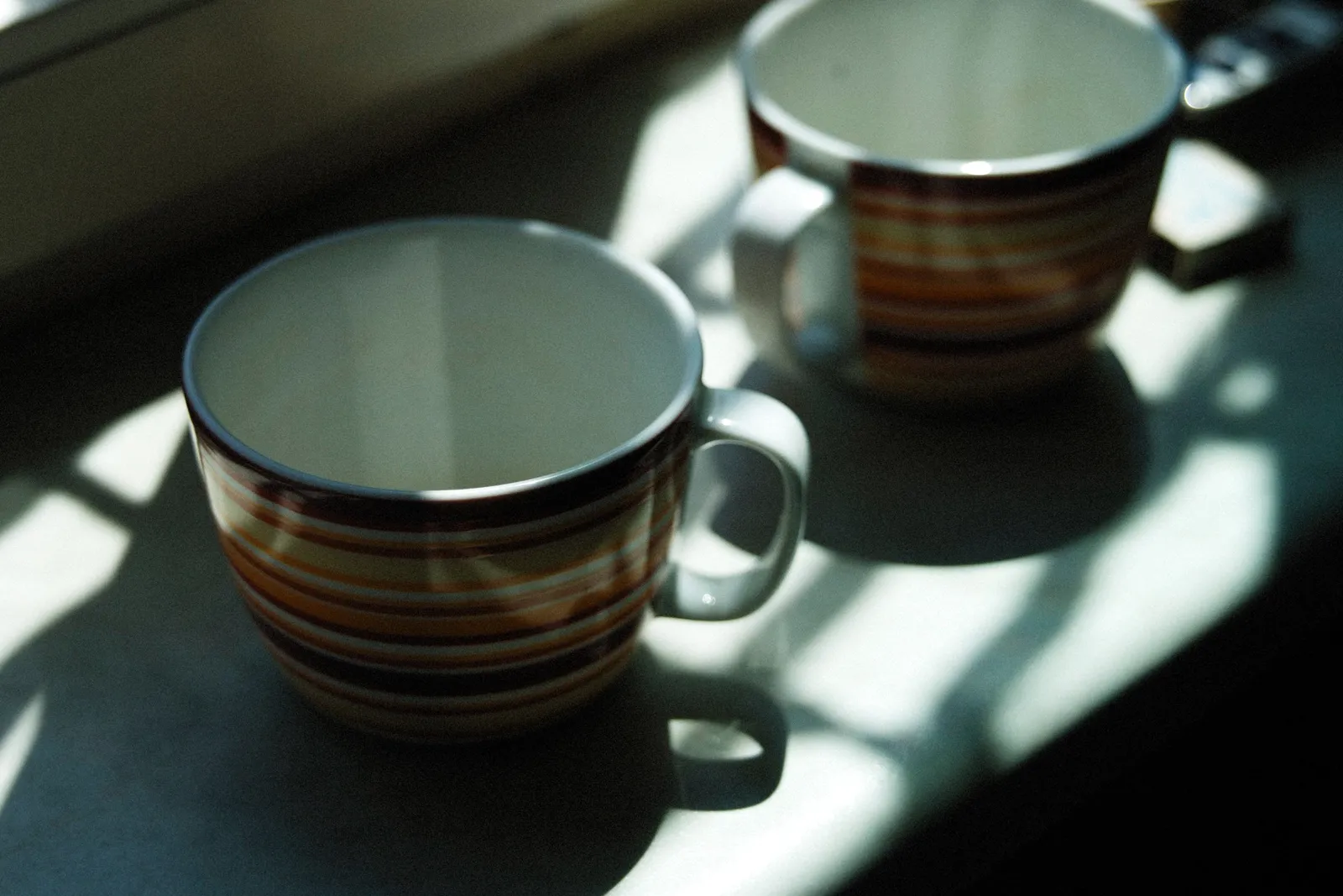 Dos tazas rayadas sobre el alféizar de una cocina, atravesadas por sol de mañana.
