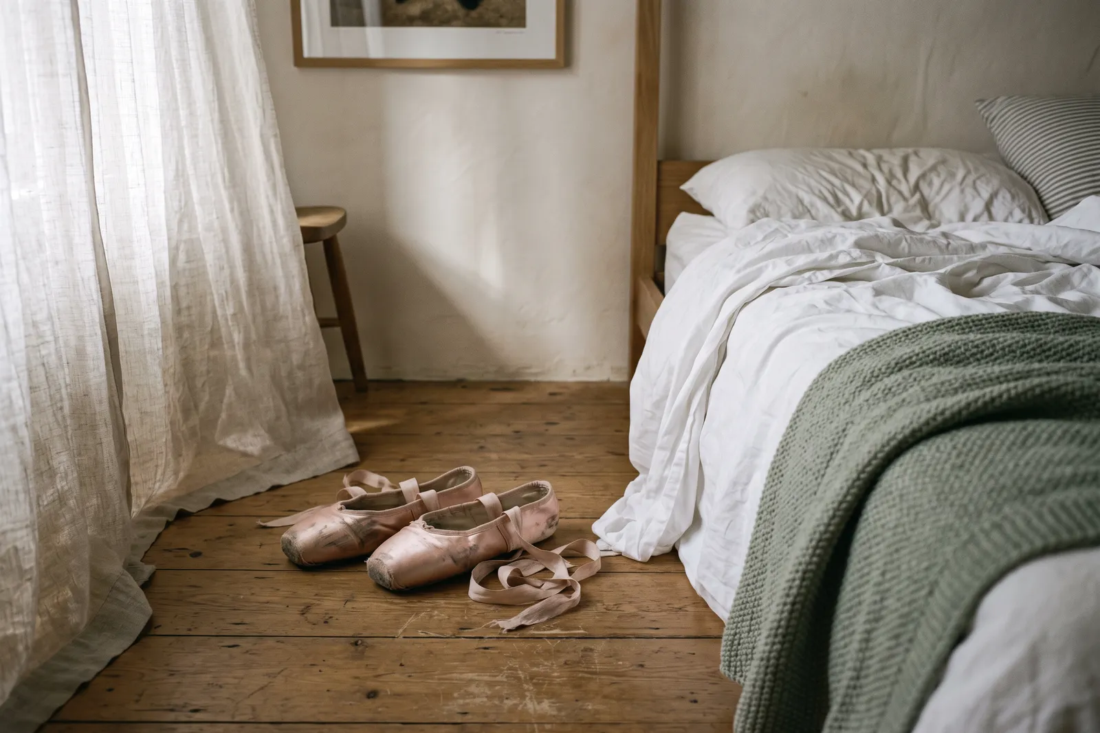 Unas zapatillas de punta con cintas sobre el piso de madera, junto a una cama deshecha con luz de mañana.