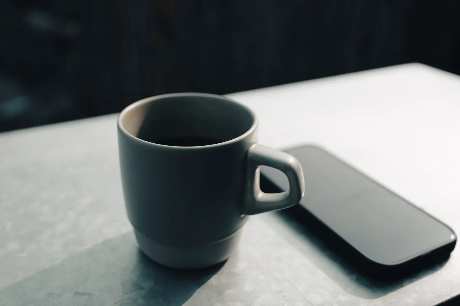 Una taza de cerámica y un teléfono sobre una mesa, con luz suave y lateral.