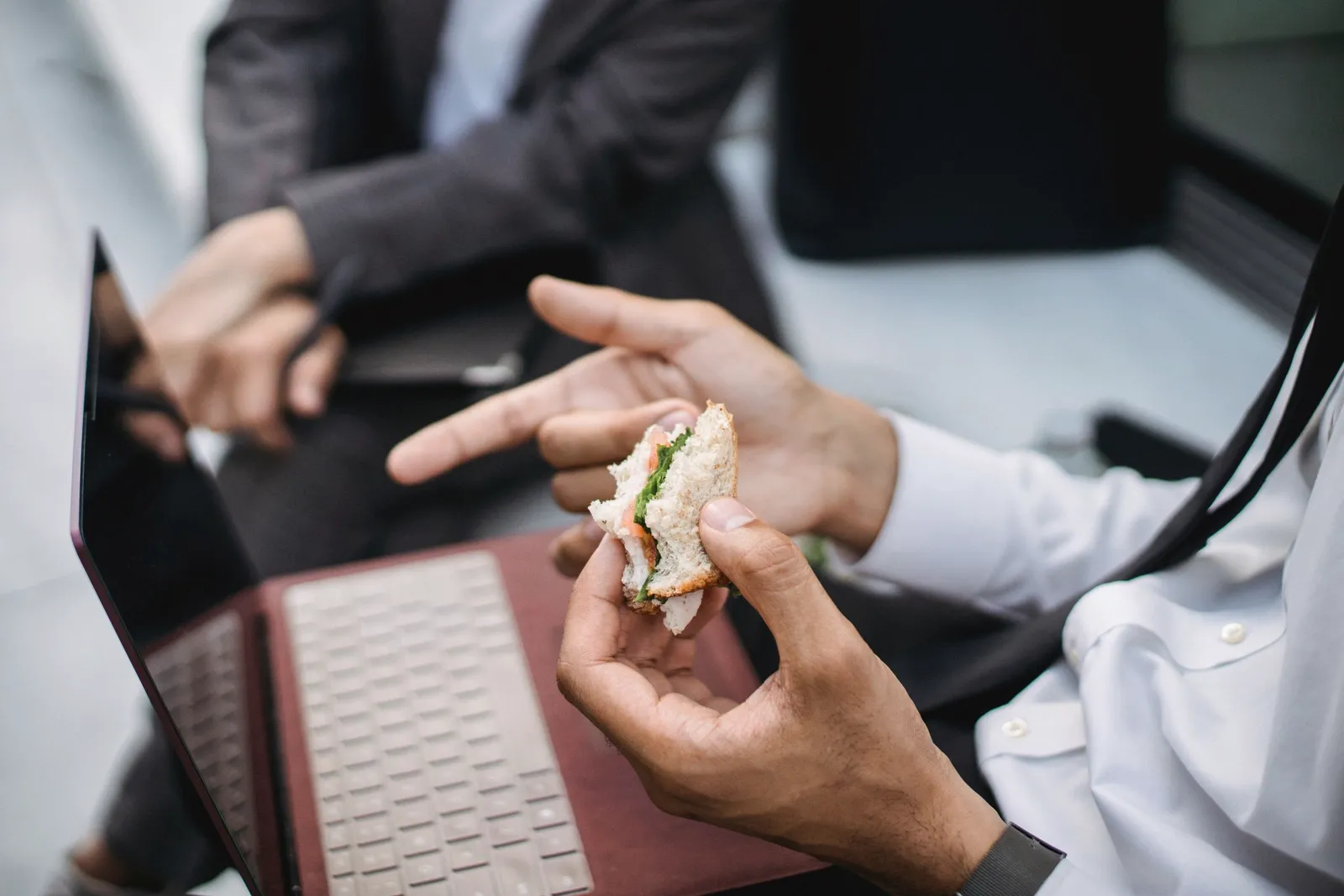 Primer plano de una mano sosteniendo un sándwich mientras la laptop sigue abierta al lado.