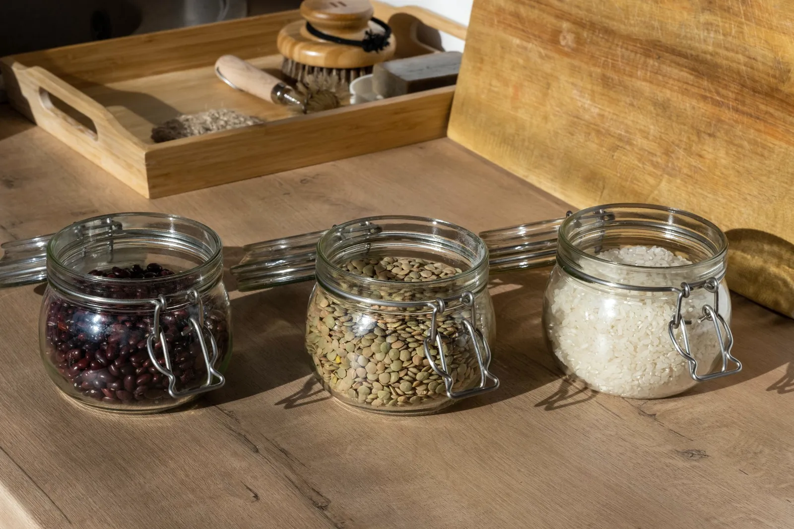 Frascos de vidrio con porotos, lentejas y arroz sobre una mesa de madera, con luz cálida.