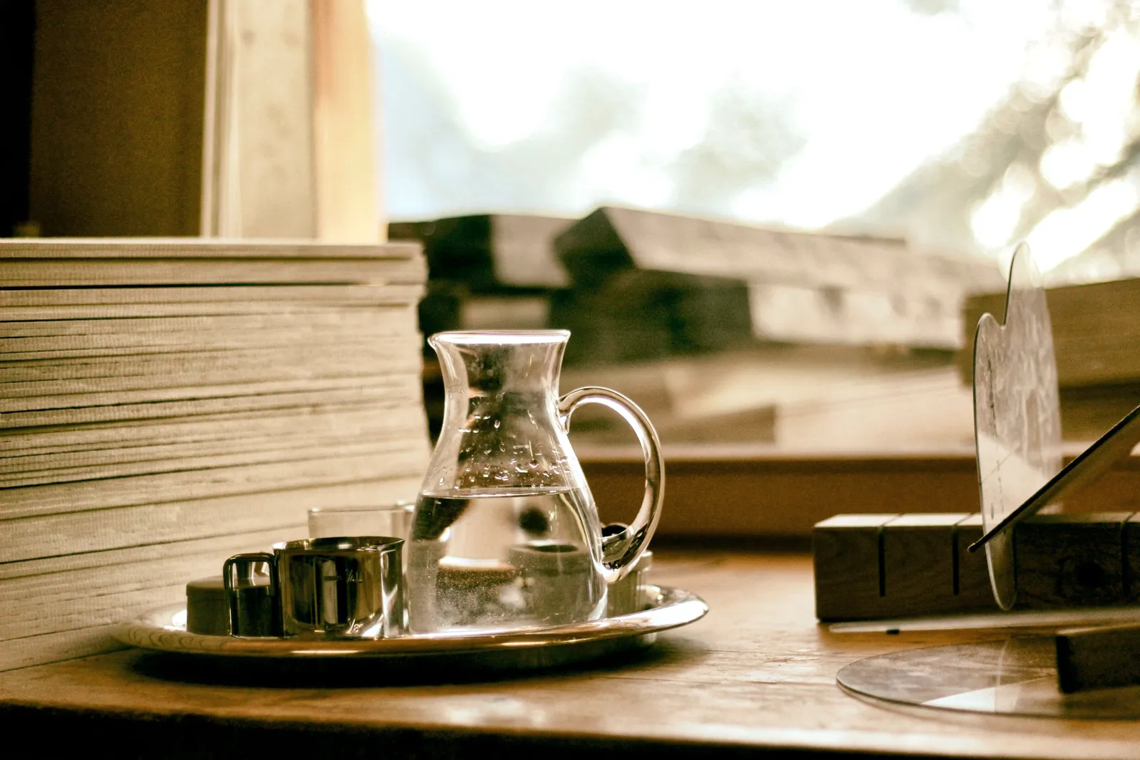 Una jarra de agua de vidrio con vasos y unos libros sobre una mesa de madera junto a la ventana.