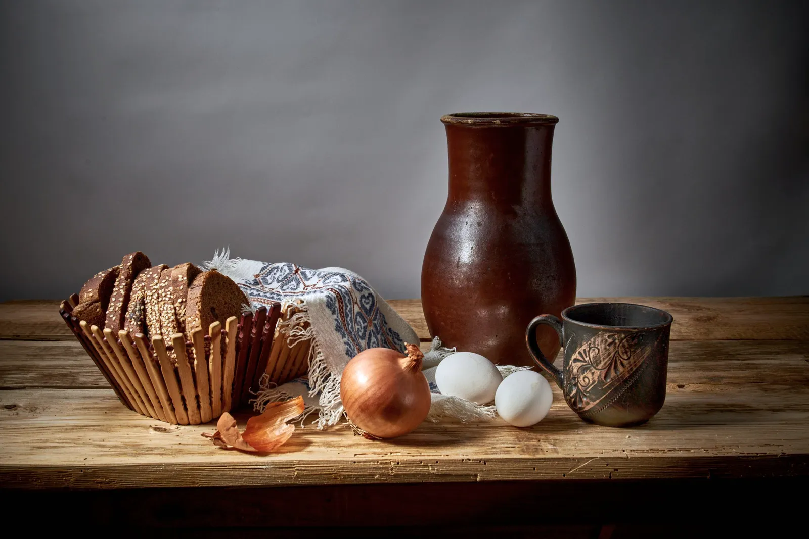 Un bodegón rústico con pan, cebollas, huevos y piezas de barro sobre una mesa de madera.