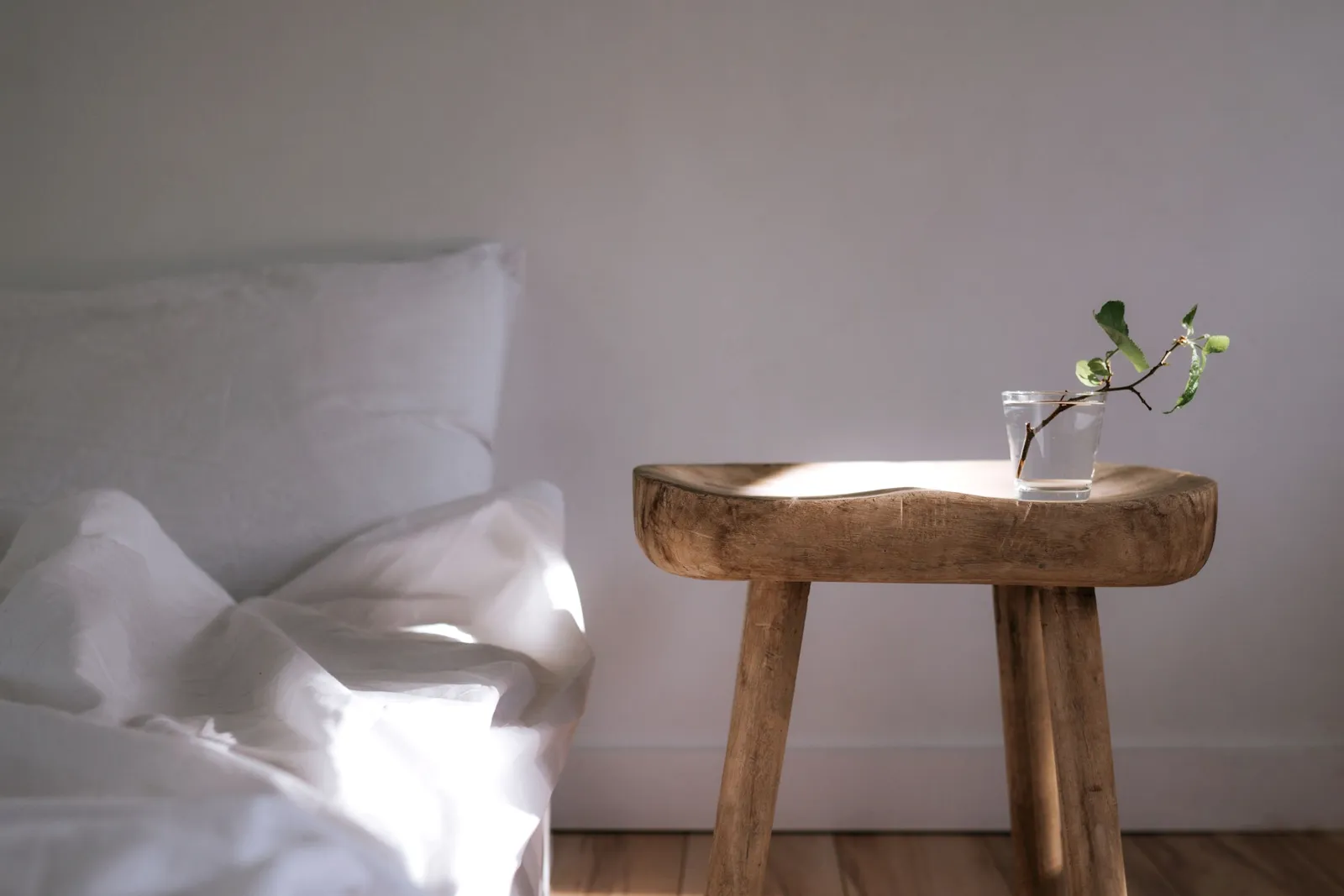 Dormitorio mínimo iluminado por el sol, con sábanas blancas y una mesa de madera con una planta.