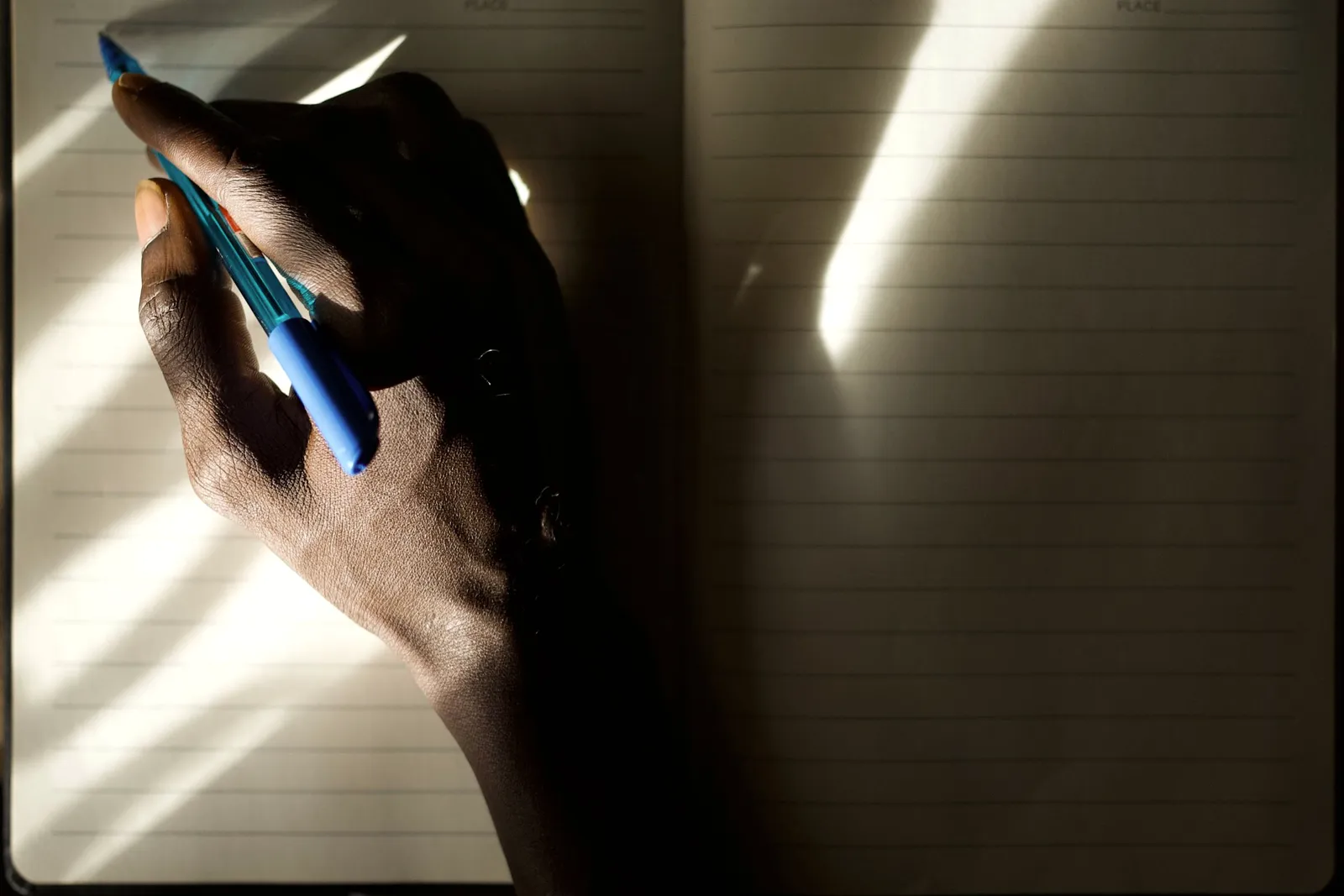 Una mano escribiendo en un cuaderno, iluminada por luz natural suave que deja sombras largas.