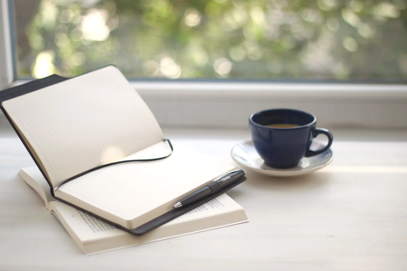 Un cuaderno abierto y un café sobre una mesa junto a una ventana con sol.