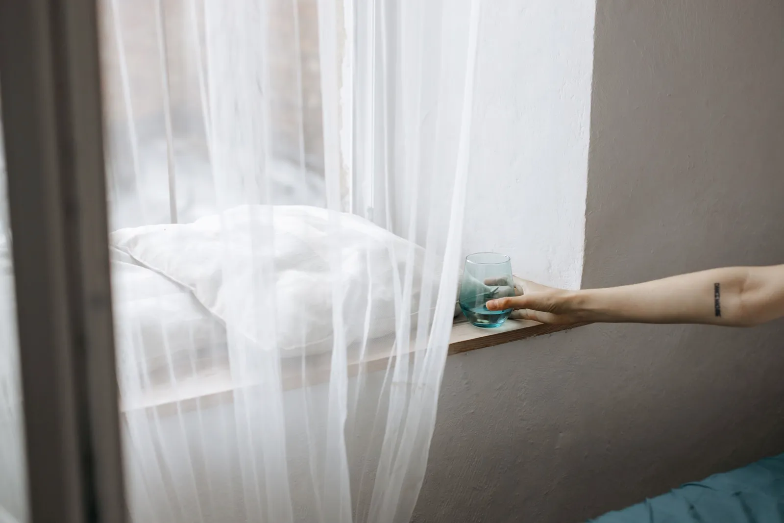 Un brazo apoyando un vaso de agua en el alféizar, con cortinas livianas y luz ambiente.