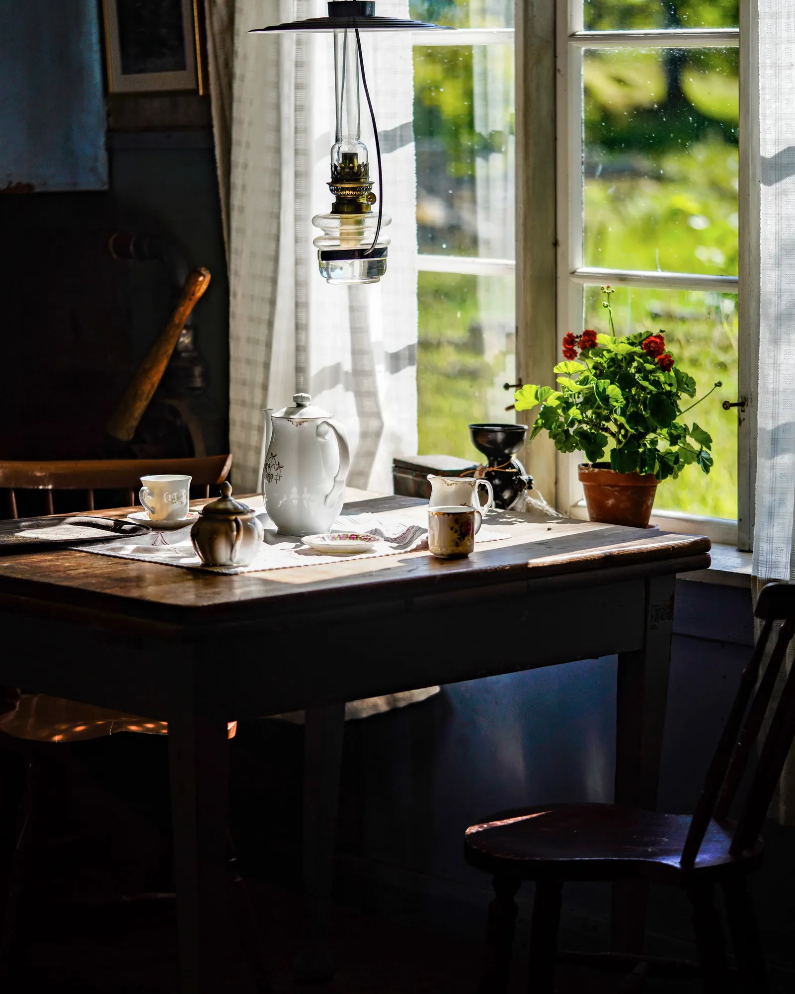 Una mesa junto a una ventana soleada con tetera, tazas y ramas verdes.