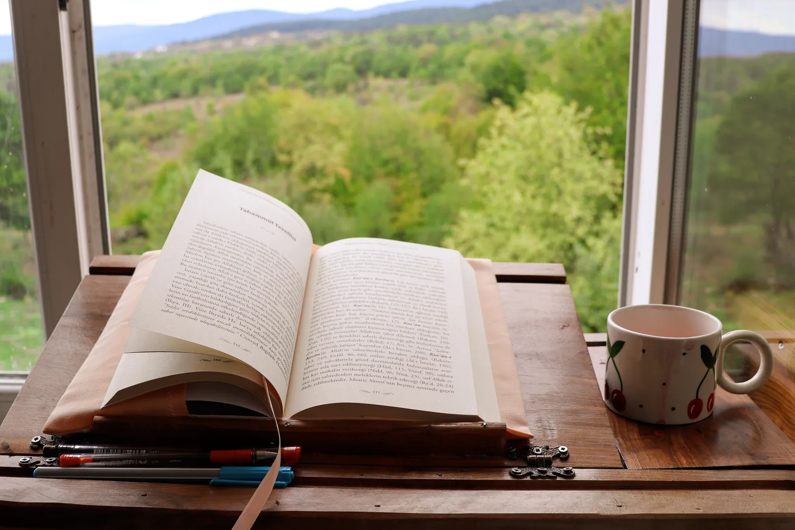 Un libro abierto y una taza de cerámica sobre una mesa junto a una ventana con vista.
