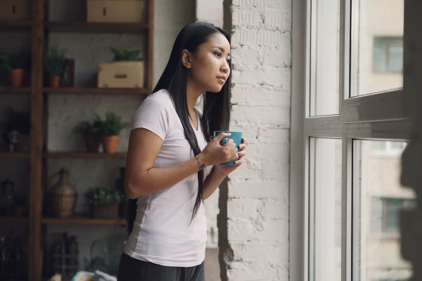 Mujer joven mirando por la ventana con una taza, en una habitación con plantas.